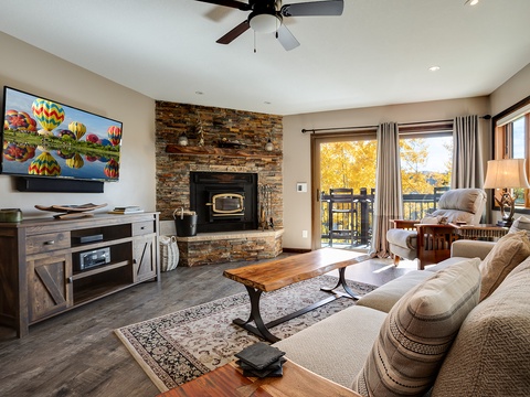 Unwind in your cozy living room featuring a stone fireplace, plush seating, and balcony access with serene views—perfect for fall evenings and movie nights.