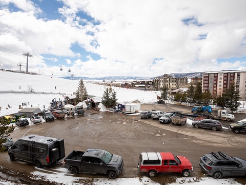 Scenic ski resort village with direct slope access, parking lot, and ski lift system connecting multi-story lodges amid snow-covered mountains under expansive skies.