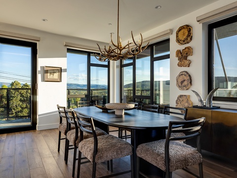 Gather around this stunning dining table with sweeping mountain and water views through floor-to-ceiling windows. The rustic antler chandelier and natural wood accents create a warm, inviting space for your memorable meals together.
