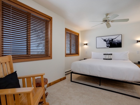 Your serene bedroom offers a crisp, welcoming bed with modern art above, warm wood touches, and ceiling fan for comfort, all framed by natural light.