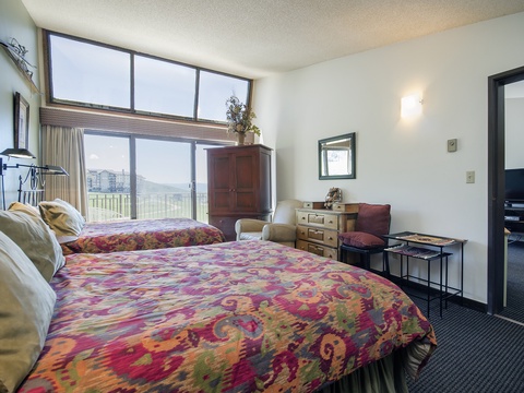 Your spacious bedroom features two plush beds with vibrant bedding, a cozy seating area, and sweeping mountain views through floor-to-ceiling windows that fill the room with natural light.