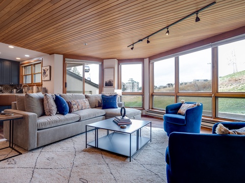 Sink into plush seating and let natural light flood your space through floor-to-ceiling windows framing mountain views. Your cozy retreat features warm wood ceilings and stylish furnishings perfect for unwinding after adventure.