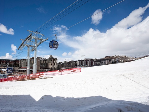 A winter ski resort with snow-covered slopes, gondola lift access, and surrounding mountain village architecture under clear blue skies.