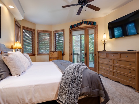 Your spacious bedroom features a plush bed, warm wood furnishings, ceiling fan, and private balcony access—perfect for relaxing after mountain adventures.