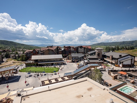 Aerial view of a mountain resort village showcasing mixed-use development with lodges, commercial buildings, recreational green space, and scenic forested mountain backdrop under summer skies.