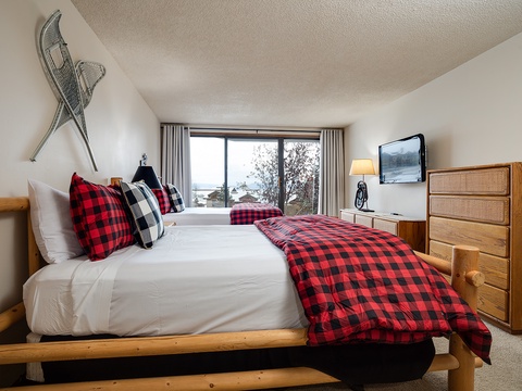 Cozy up in this charming bedroom with rustic wood furnishings, plaid accents, and a serene lake view right outside your window—your perfect mountain escape.