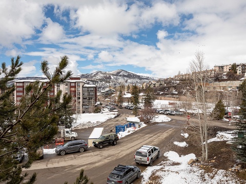 A scenic winter setting nestled in a mountain community, with snow-dusted peaks framing residential buildings and parking areas amid evergreen trees and clear mountain air.