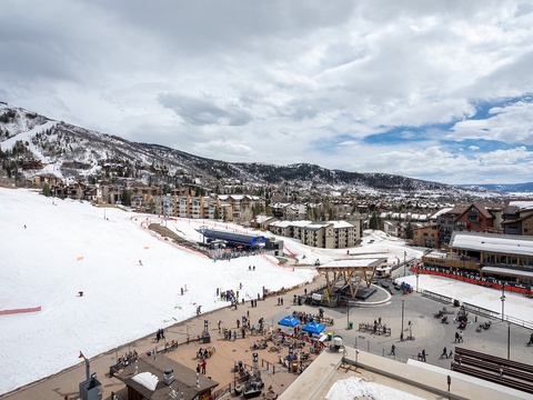 Aerial view of a mountain resort village nestled against snow-covered slopes, featuring ski facilities, lodges, and a bustling base area with shops and restaurants.