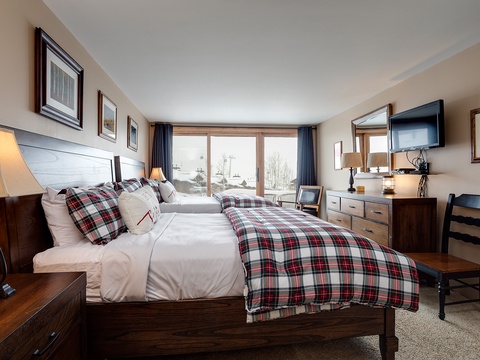 Relax in your cozy bedroom with plaid bedding, warm lighting, and a flat-screen TV. The large windows frame serene winter views—perfect for unwinding after a day of adventure.