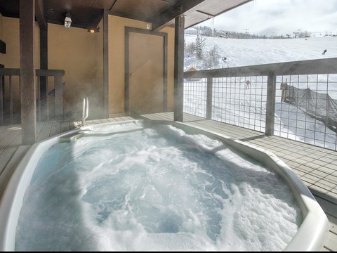 A bubbling hot tub on a covered terrace offers stunning views of snow-covered ski slopes and mountain scenery, perfect for relaxing après-ski moments.