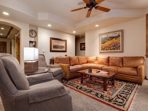 Sink into plush seating and unwind in your spacious living room, where a cozy sectional and armchairs invite relaxation beneath a gently circulating ceiling fan and warm ambient lighting.