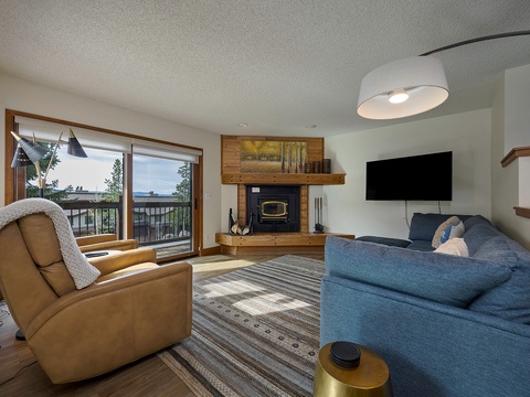 Settle into your cozy living room where sunlight streams through sliding glass doors, revealing forest and lake views. Unwind by the wood-burning fireplace or watch shows on the flat-screen TV.