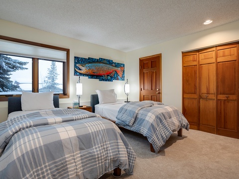 Two plush beds adorned with soft plaid linens invite you to rest after a day of exploring, while evergreens framed by the window and a vibrant trout artwork set a cozy, mountain retreat mood.