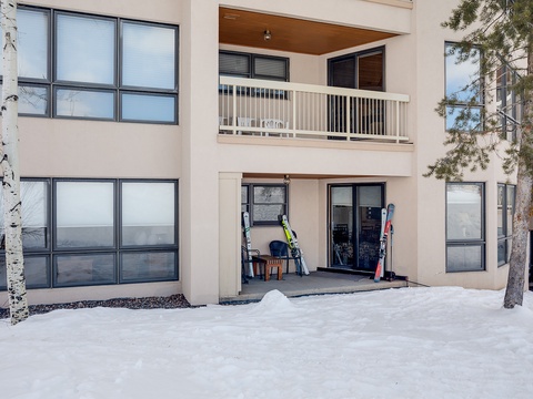 Modern mountain property entrance with ski storage at ground level and private balcony above, surrounded by snow-covered grounds and natural pine trees.