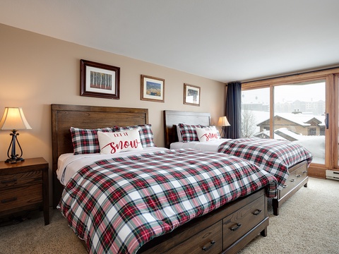 Cozy up in this alpine bedroom where plaid bedding meets breathtaking snowy mountain views through expansive windows—your perfect winter retreat awaits.
