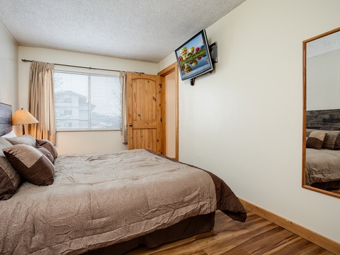 Your cozy bedroom features a plush queen bed, warm wood accents, a wall-mounted TV for entertainment, and natural light streaming through the window—perfect for peaceful mornings and restful nights.