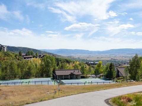 Mountain resort property featuring tennis courts nestled in a scenic alpine valley with expansive views of forested hillsides and distant peaks.