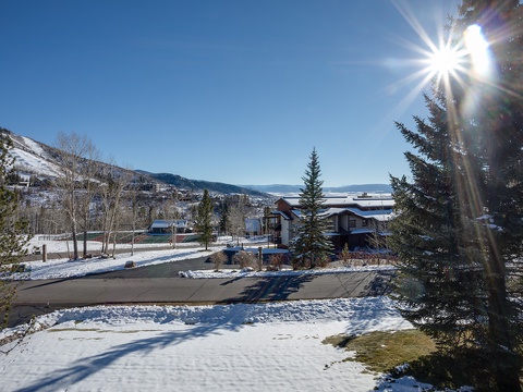 A picturesque winter setting with snow-covered mountains, evergreen trees, and a quiet residential street under brilliant blue skies and sunshine.