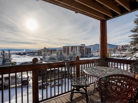 A picturesque winter mountain resort scene surrounded by snow-dusted buildings and alpine peaks, framed by a covered wooden balcony with seating for outdoor relaxation.