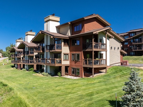 Multi-level alpine-style condominiums with private balconies, nestled in a scenic mountain setting surrounded by manicured lawns and evergreen trees.