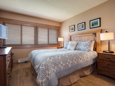 Your peaceful retreat features a plush bed with soft linens and warm wooden accents. Gentle light filters through the blinds as you relax surrounded by cozy furnishings and tasteful artwork.