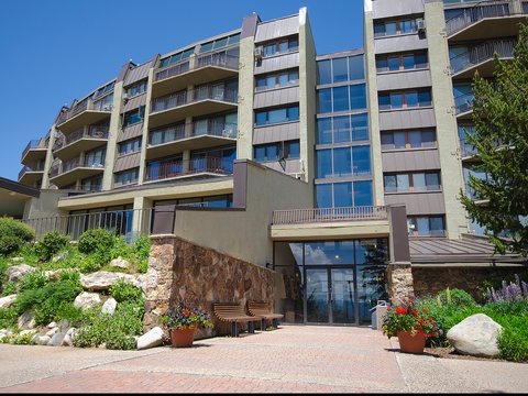A modern vacation rental property with multiple balconies nestled in a beautifully landscaped garden setting, offering a welcoming entrance amid lush greenery and colorful blooms.