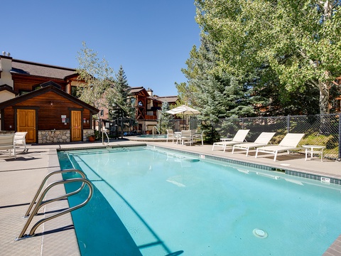The property features a seasonal outdoor pool surrounded by mountain pines and deck chairs, with a wooden cabin and alpine-style buildings visible around the courtyard.