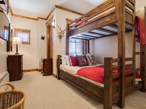 Your cozy alpine-style bedroom features a sturdy bunk bed with plaid accents, warm wood trim, and charming rustic touches like the antler mount and wicker seating—perfect for restful mountain nights.