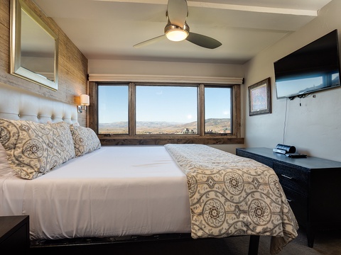 Wake up to sweeping vistas framed by rustic wood windows. Your cozy retreat features a plush tufted headboard, modern ceiling fan, and flat-screen TV for unwinding after a day exploring the landscape.