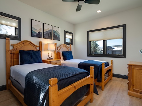 Your cozy alpine retreat features two twin beds with plush bedding, warm wood accents, and peaceful mountain views through large windows inviting natural light into the room.