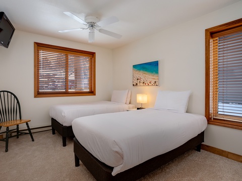 Two cozy beds with crisp white linens invite you to unwind after a day of adventure. Natural light filters through warm wood blinds, while a beach scene reminds you of the coast nearby.