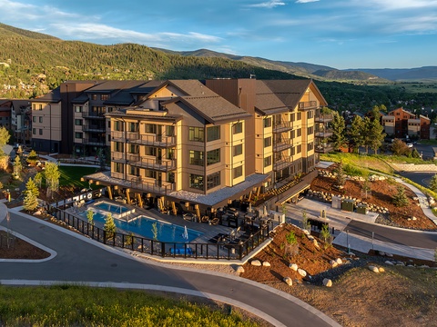 An aerial view captures the property at the base of forested mountains, featuring outdoor pool areas and a multi-story main building nestled within a scenic mountain community. The surrounding hillsides showcase the natural landscape of the area.