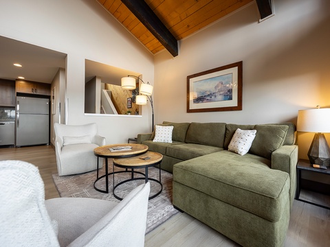 Relax in your open-concept living space featuring a plush sectional sofa, warm vaulted wood ceilings with exposed beams, and a thoughtfully appointed kitchen waiting for your culinary adventures.