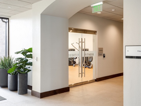 A modern fitness center awaits behind sleek glass doors, with equipment visible and ready for your workout sessions. Fresh greenery adds a welcoming touch to the entrance.