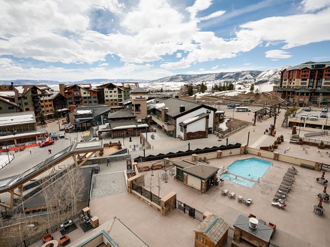 Elevated view showcasing the property's surrounding alpine village, featuring a pool area, mountain-facing buildings, and nearby ski resort amenities set against a winter landscape.
