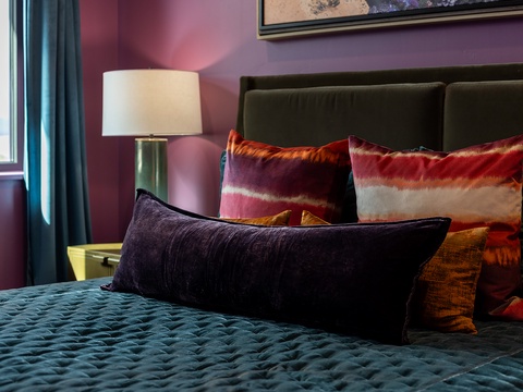 Sink into luxurious comfort with plush textured pillows and a quilted teal bedspread, all framed by bold purple walls and soft natural light from your window.