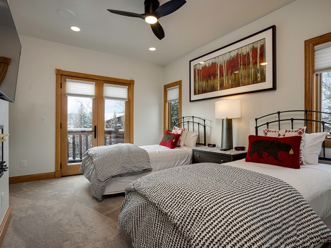 Your cozy bedroom features two plush beds with crisp linens, warm lighting from bedside lamps, and a ceiling fan for comfort. Wake to snowy mountain views through wood-framed windows.