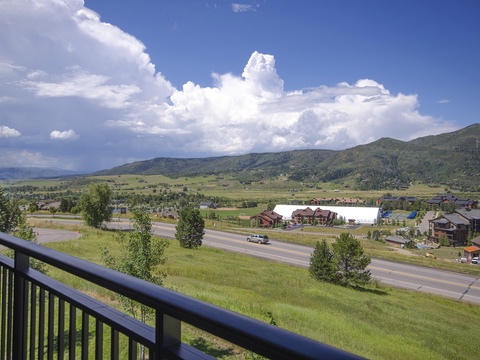 Mountain valley framed by rolling green hillsides, scattered homes, and an open highway, all under a vivid blue sky with billowing summer clouds.
