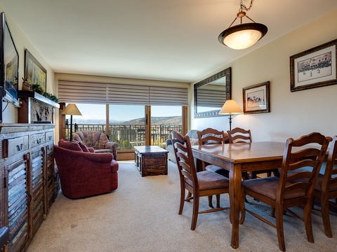 Your open living and dining area frames stunning mountain views through large balcony doors, with comfortable seating, a full dining table for six, and cozy ambient lighting throughout.