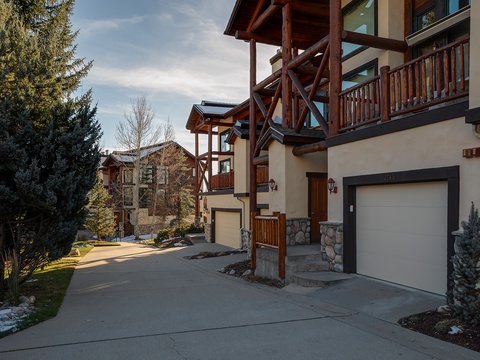 The property sits in a peaceful mountain neighborhood, featuring rustic timber balconies and convenient garage access, with natural landscaping and neighboring chalets visible along the quiet street.