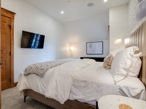 Cozy up under plush bedding with a wall-mounted TV and modern lighting. Your peaceful retreat features a textured accent wall and warm, inviting touches to help you unwind.