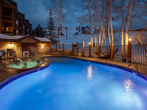 Mountain resort featuring an illuminated outdoor pool and steaming hot tub, nestled among snow-dusted aspen trees with ski slopes visible in the background at twilight.