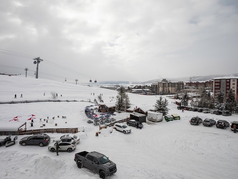 Aerial view of a snowy ski resort with slopes, lodges, parking areas, and gondola lifts set against a mountainous winter landscape.