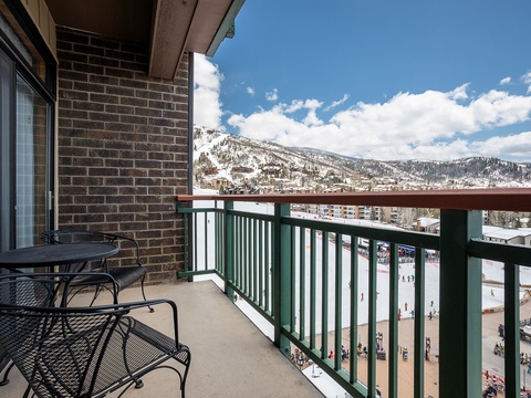 A winter vacation rental balcony overlooking an ice skating rink and snow-capped mountain slopes, with outdoor seating for watching seasonal activities below.