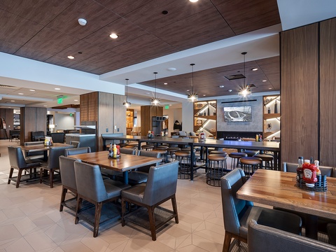 A contemporary communal space invites you to gather and dine with fellow travelers, featuring warm wood tones, modern lighting, and a cozy fireplace visible in the distance.