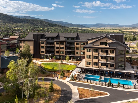 An aerial view of this modern mountain resort property, surrounded by rolling foothills and distant peaks. The well-appointed grounds feature lush lawns, outdoor pools, and scenic valley vistas stretching beyond the property.