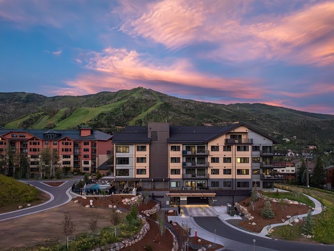 A resort property with modern multi-story buildings and ski slopes nestled at the foot of green mountains beneath a colorful sunset sky.