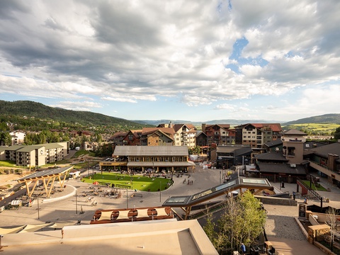 An aerial view showcasing a mountain resort village surrounded by forested hills and distant peaks, with communal spaces and lodging buildings clustered in the valley below.