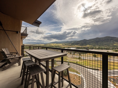 An elevated balcony with bar-height seating offers an unobstructed view of rolling mountains and sweeping valley terrain, framed by dramatic clouds overhead.