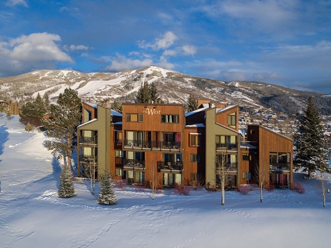The West condominium building sits nestled in snow with ski slopes and mountain peaks creating a stunning winter backdrop in this popular resort area.